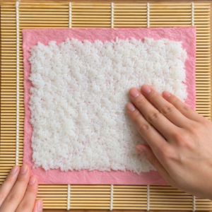 Spreading a thin layer of sushi rice over a pink soy paper sheet on a bamboo mat
